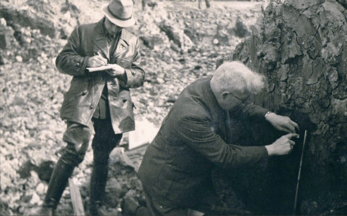 Pfaffenberg (rechts) vor einem Profil knieend. Ein Mitarbeiter notiert seine Angaben.